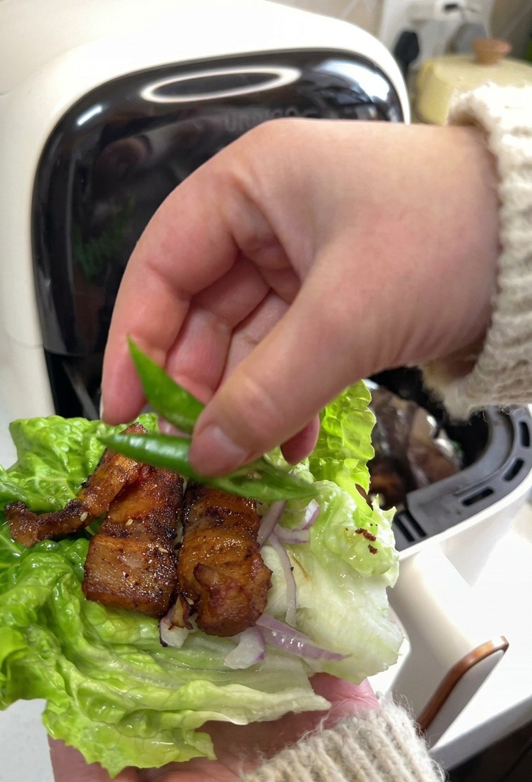 A hand placing two bright green chili slices onto a lettuce leaf filled with roasted pork belly and sliced red onions.