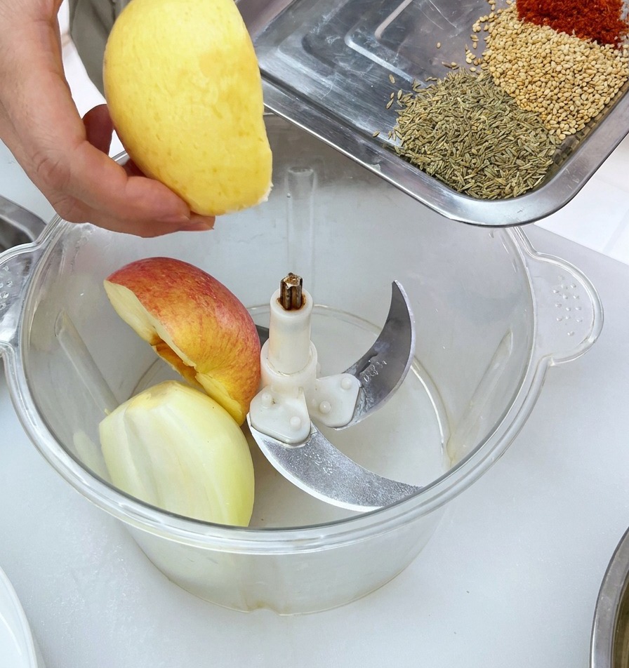 Pieces of fresh fruit being placed into a food processor bowl with a tray of spices ready to be added.