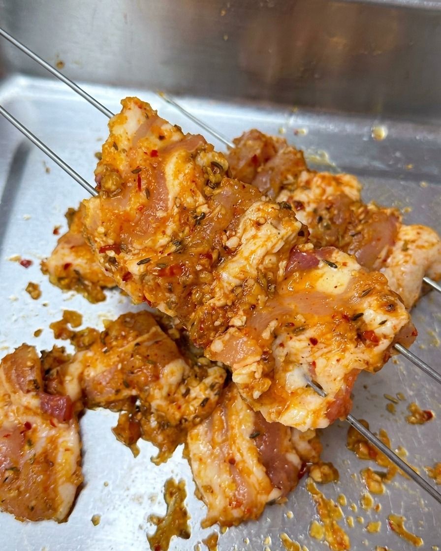 Marinated pork belly slices being pierced by two thin metal skewers over a metal prep tray.