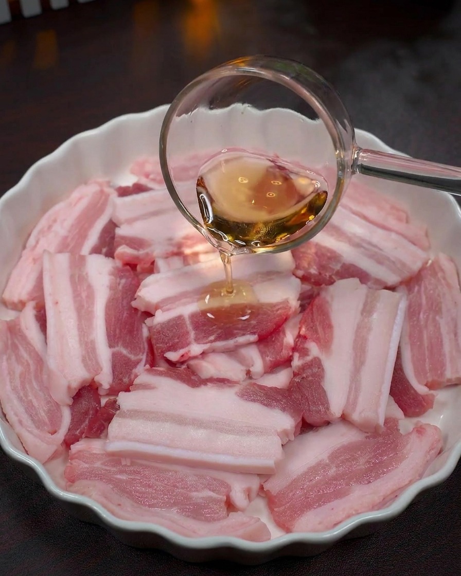 Clear liquid being poured from a glass ladle onto raw pork belly slices in a white bowl.