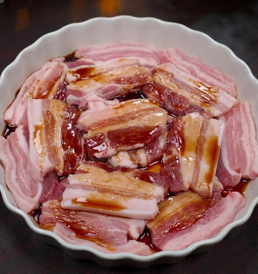 Slices of raw pork belly in a white scalloped dish being coated with a dark savory marinade.