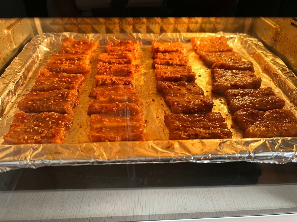 Slices of marinated pork belly roasting on a foil-lined tray inside an oven with warm interior lighting.