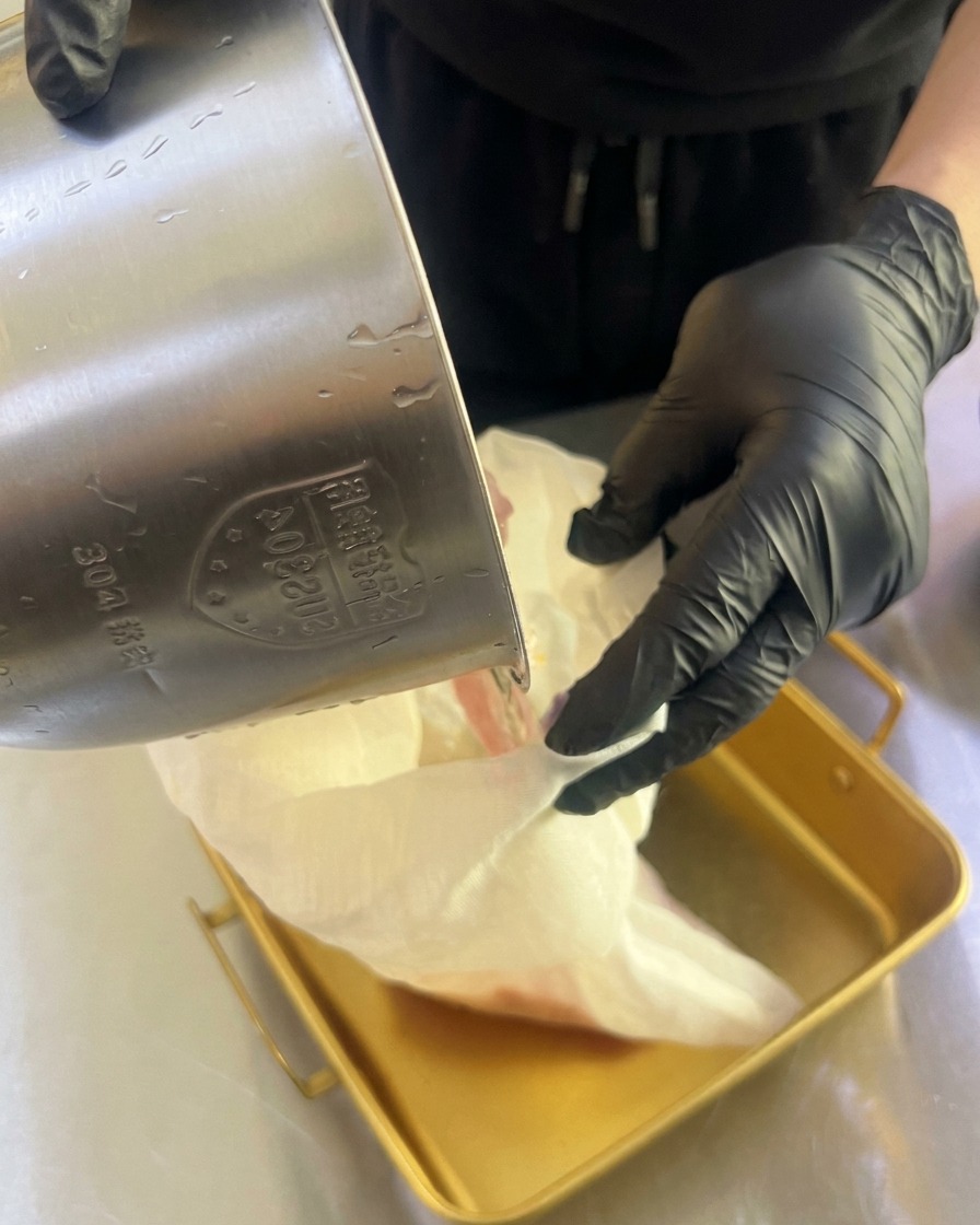 Gloved hands squeezing a white cloth bag to drip fresh fruit juice into a shallow gold metal tray.