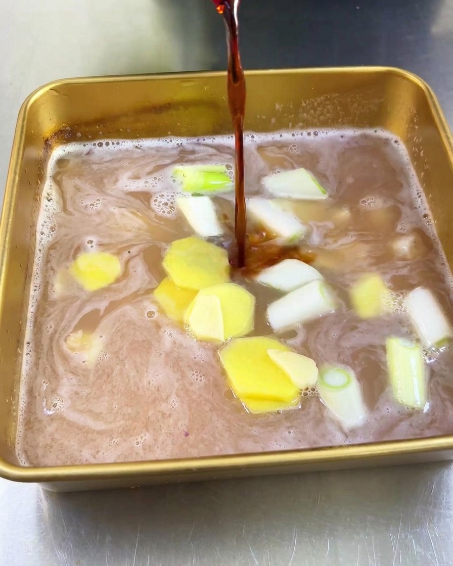 A stream of dark soy sauce being poured into a gold tray filled with light-colored fruit juice, ginger slices, and chopped green onions.