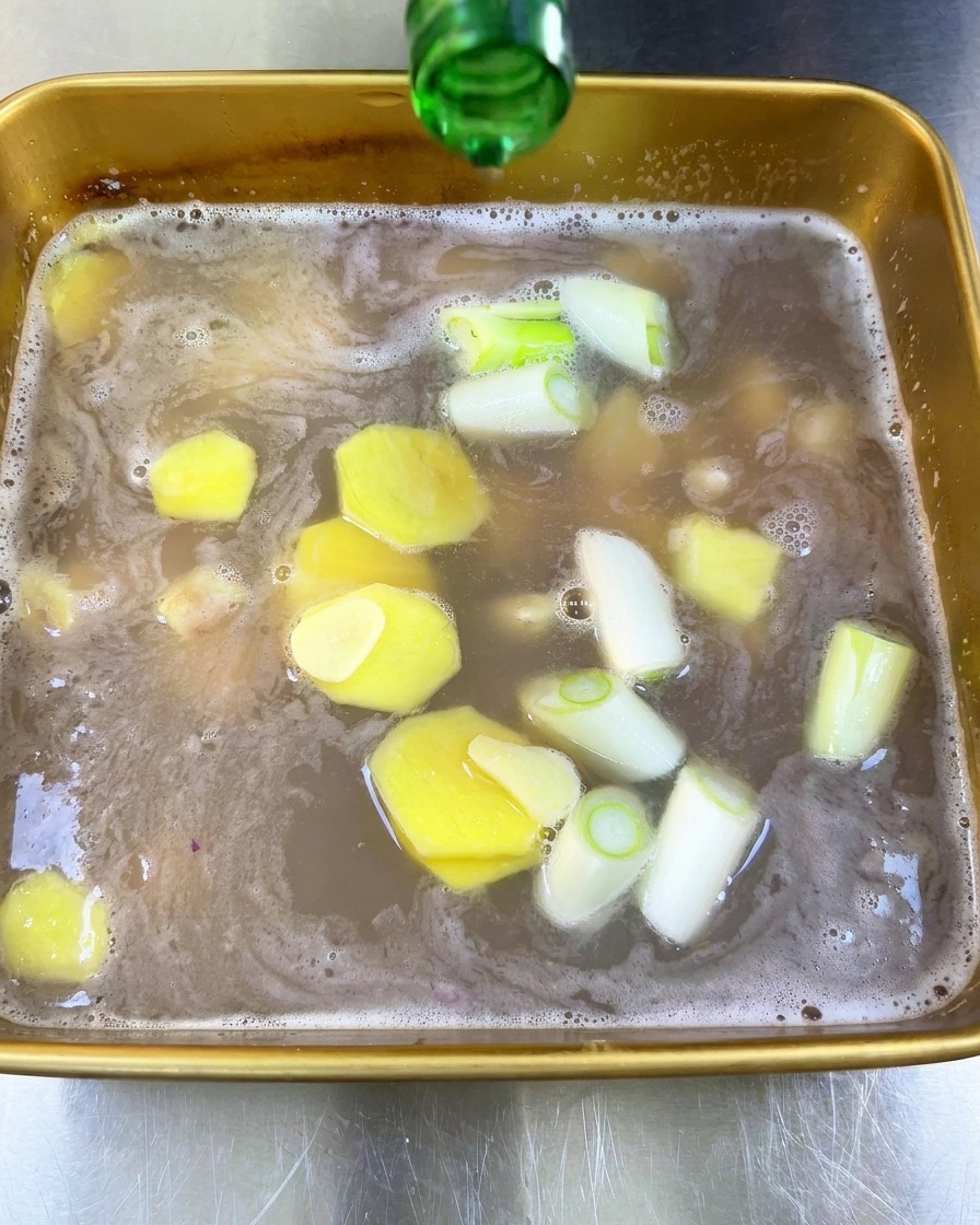 Clear rice wine being poured from the neck of a green glass bottle into a tray of liquid marinade containing ginger and scallions.