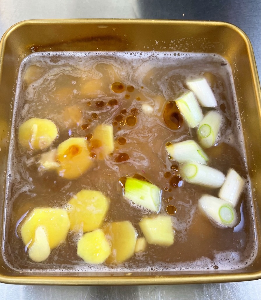 A top-down view of a golden-brown soy marinade in a square tray, featuring slices of ginger, cut green onions, and dark droplets of sesame oil floating on the surface.