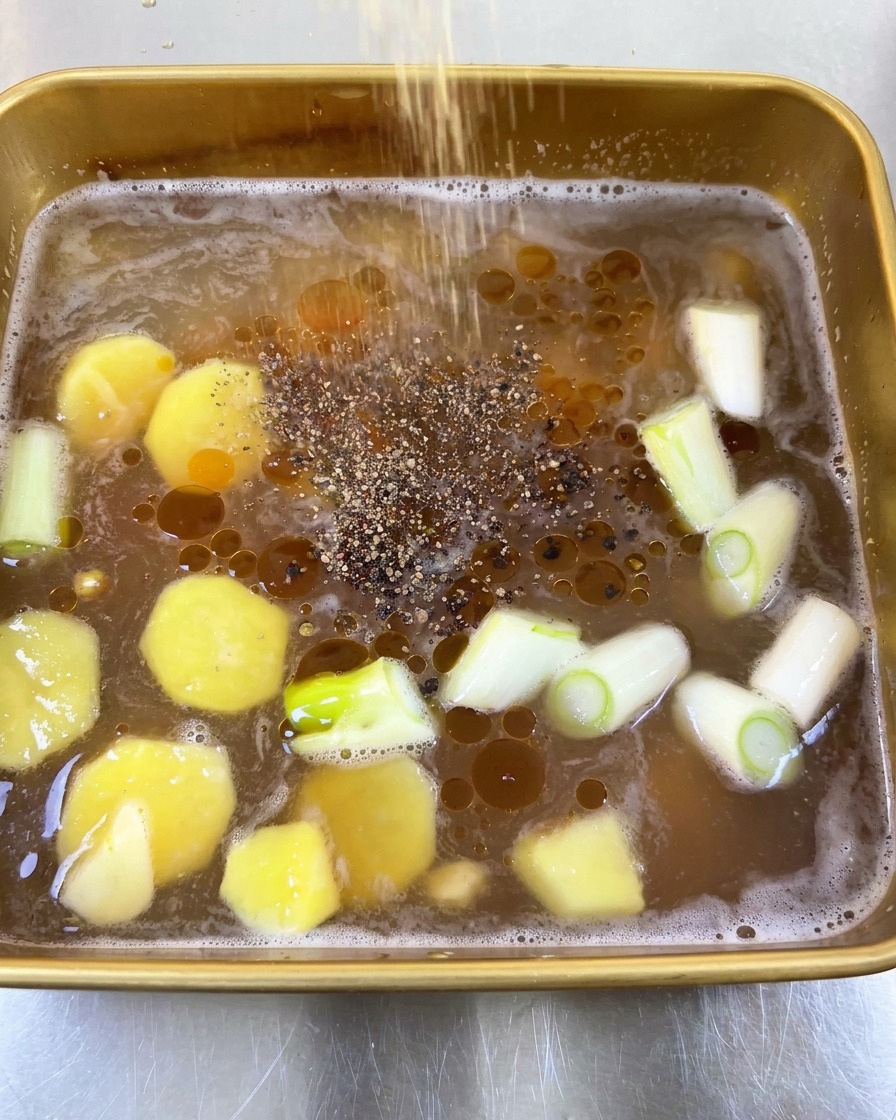 Ground black pepper being sprinkled over a liquid marinade featuring oil droplets, sliced ginger, and green onions.