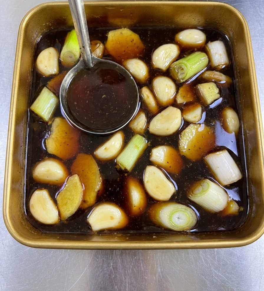 A metal ladle stirring a dark brown marinade filled with ginger and green onions in a gold square tray