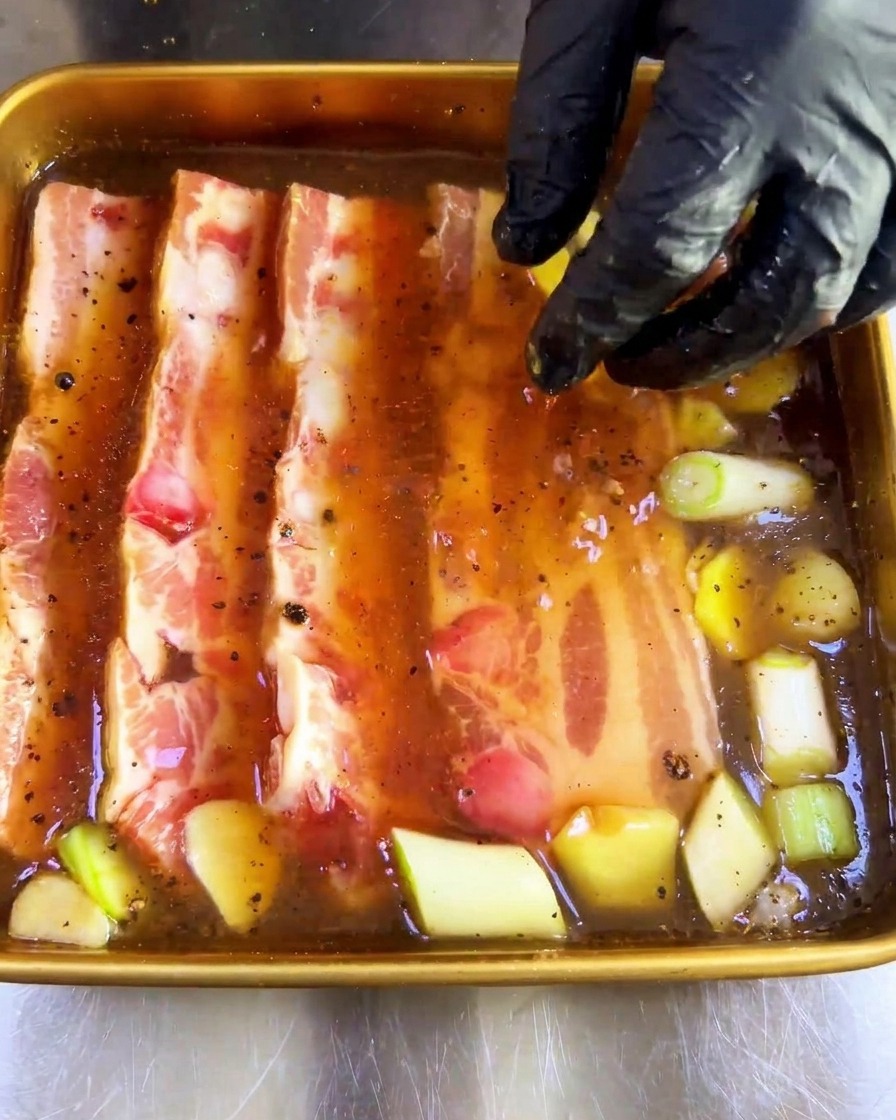 A gloved hand pressing thick, raw strips of pork belly into a tray of dark brown marinade with aromatics.