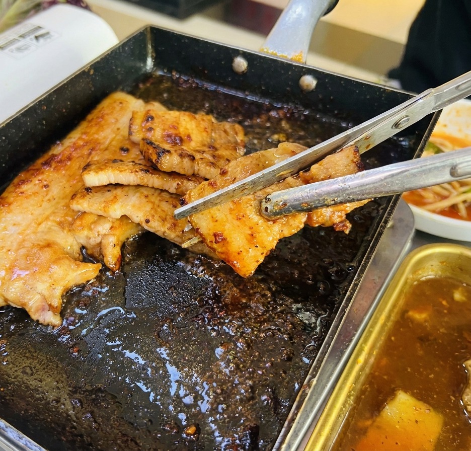 A close-up shot of thick pork belly slices being cut with metal tongs and scissors on a black square grill pan, with the meat showing a deep brown, cooked exterior.
