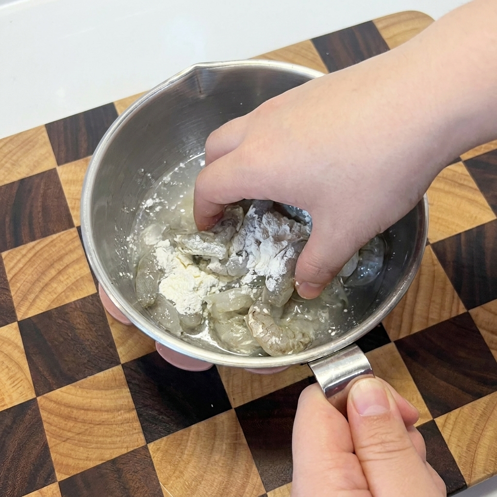 Raw shrimp being mixed with white cornstarch by hand in a metal mixing bowl.