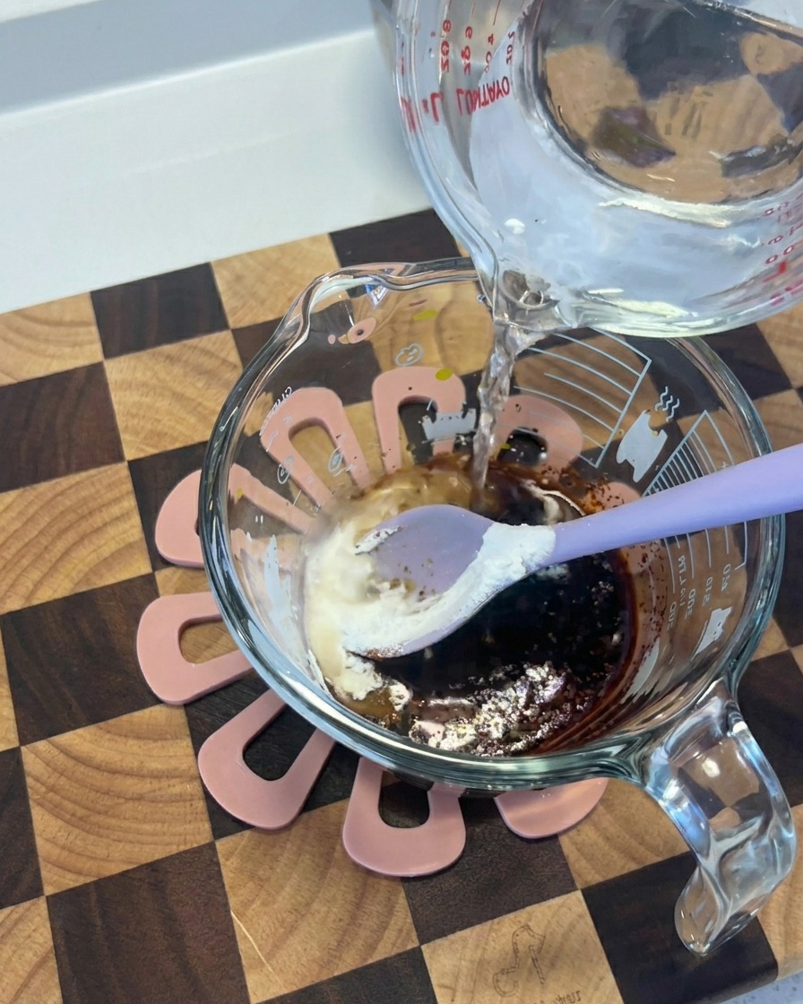 Water being poured from a glass container into another cup containing a dark mixture of soy sauce and spices.