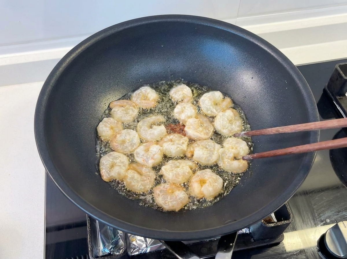 Several shrimp being stir-fried with wooden chopsticks in a black wok with shimmering oil.