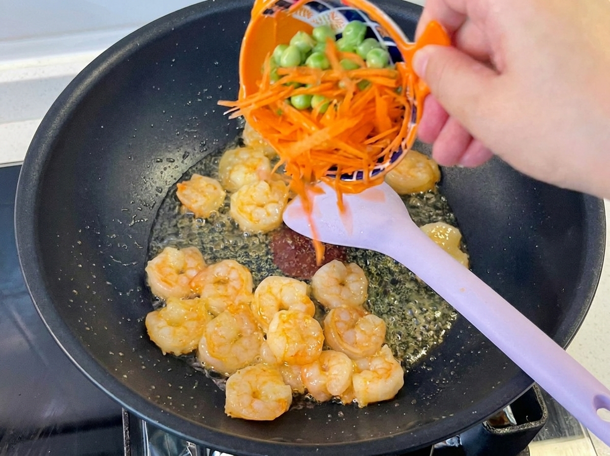 A hand pouring a mixture of bright orange shredded carrots and small green peas from a patterned orange bowl into a wok containing cooked shrimp and a purple spatula.