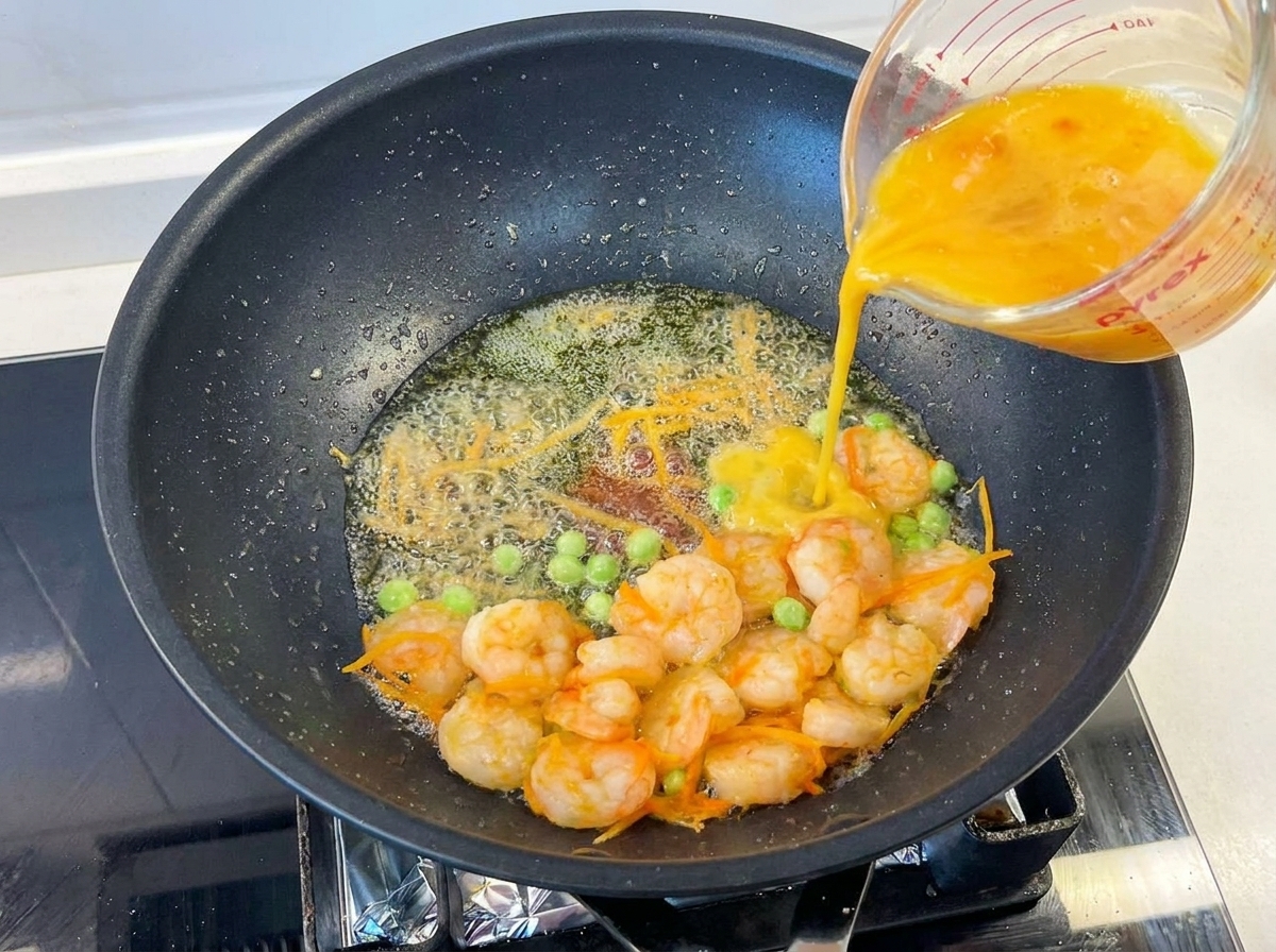 Yellow beaten eggs being poured from a glass measuring cup into a hot pan containing cooked shrimp, green peas, and shredded carrots.