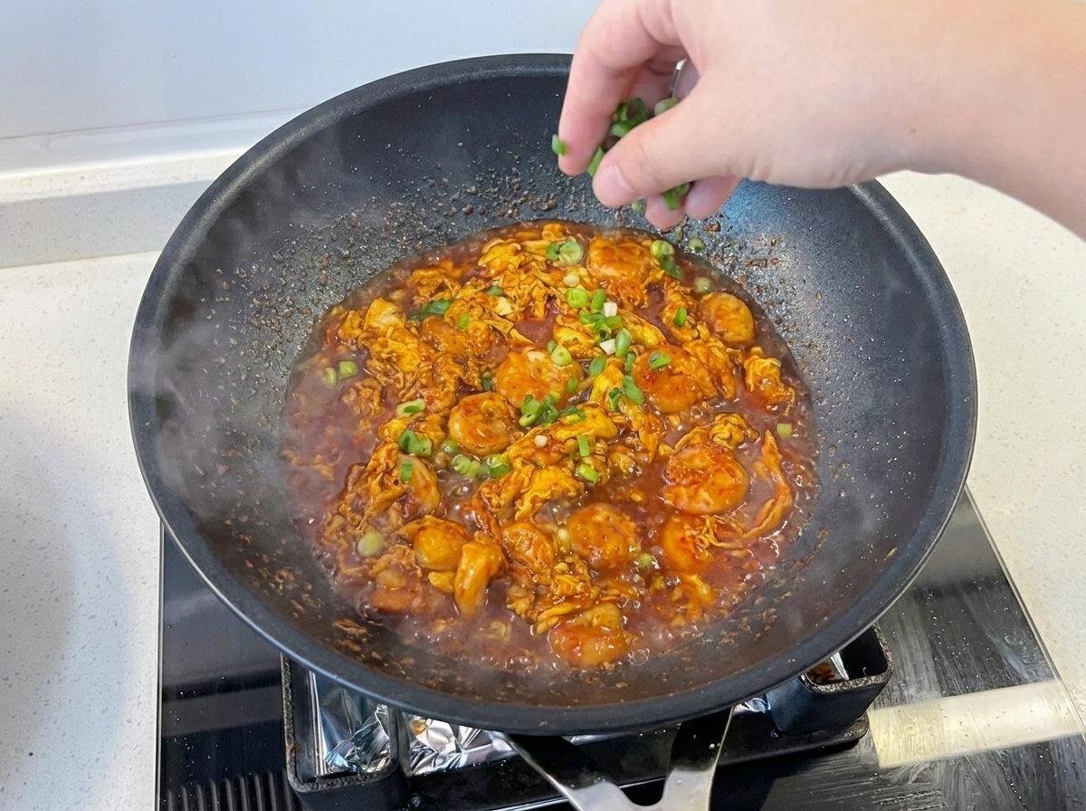 A hand sprinkling freshly chopped green onions over a vibrant, saucy shrimp and scrambled egg dish simmering in a black pan.