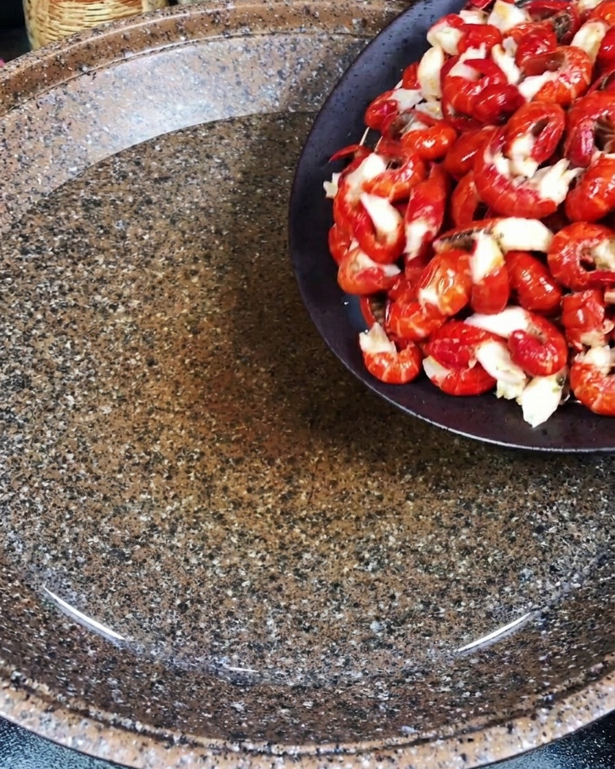 Raw red crawfish tails being poured from a dark bowl into a large pot of water containing pieces of fresh ginger.