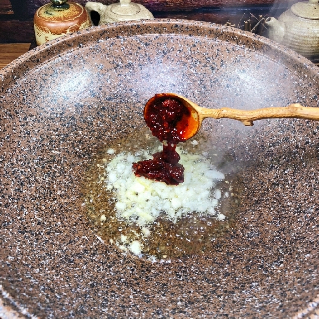 A wooden spoon adding a dollop of thick, dark red chili bean paste into a wok with sautéed minced aromatics.