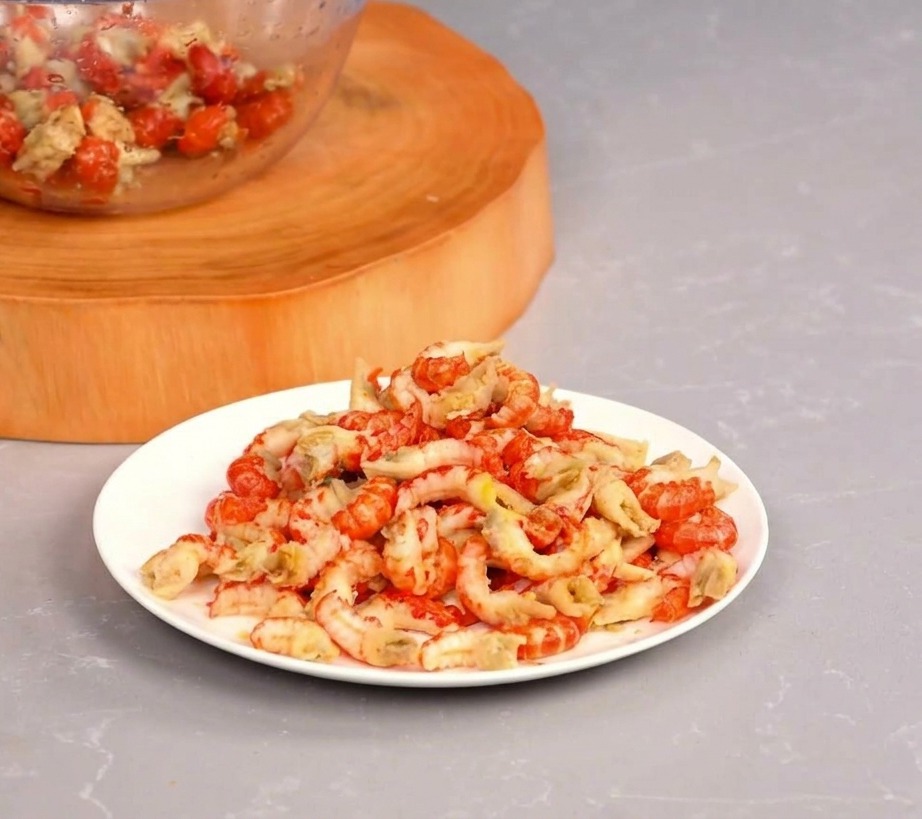 A white plate piled high with peeled red crayfish tails sitting next to a glass bowl on a wooden cutting board.