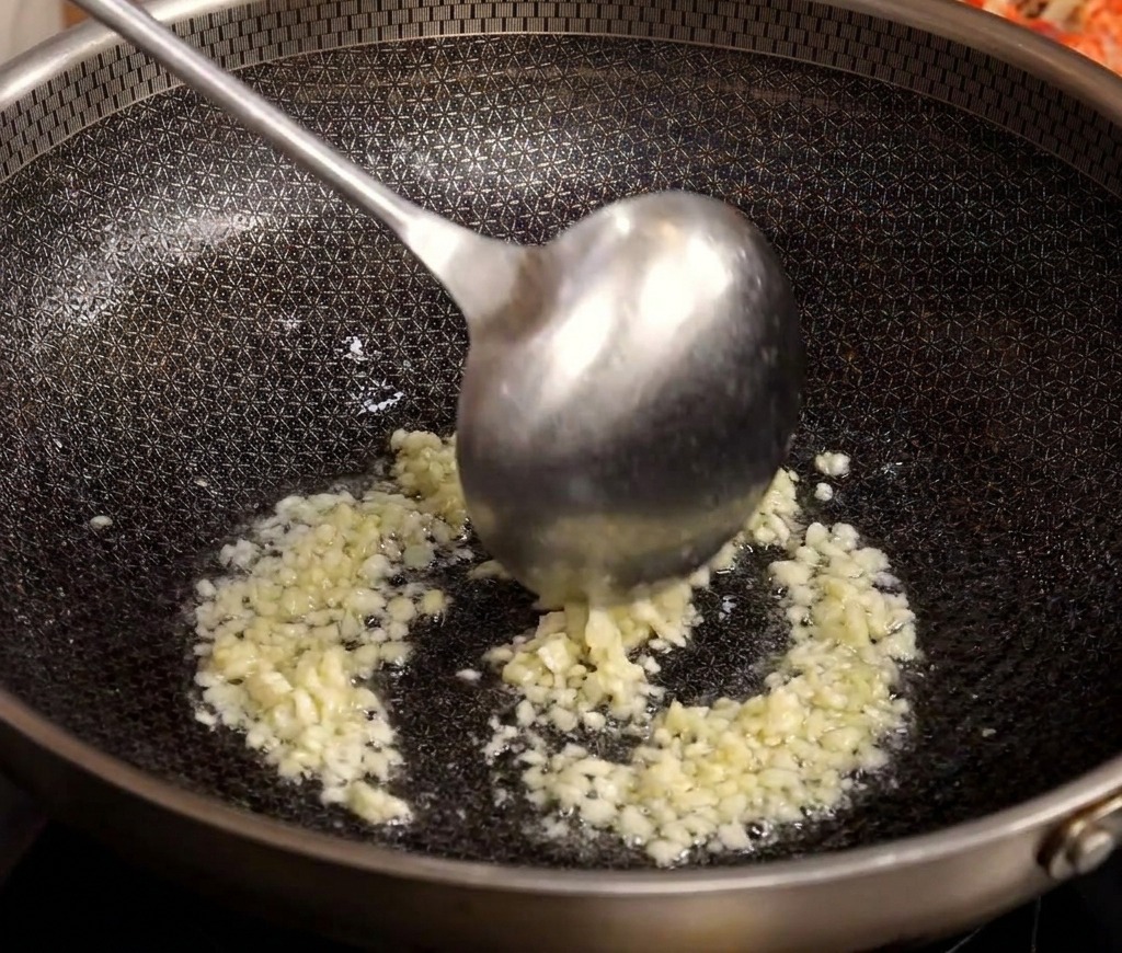 A metal ladle stirring minced garlic as it sizzles and browns in a hot wok with oil.