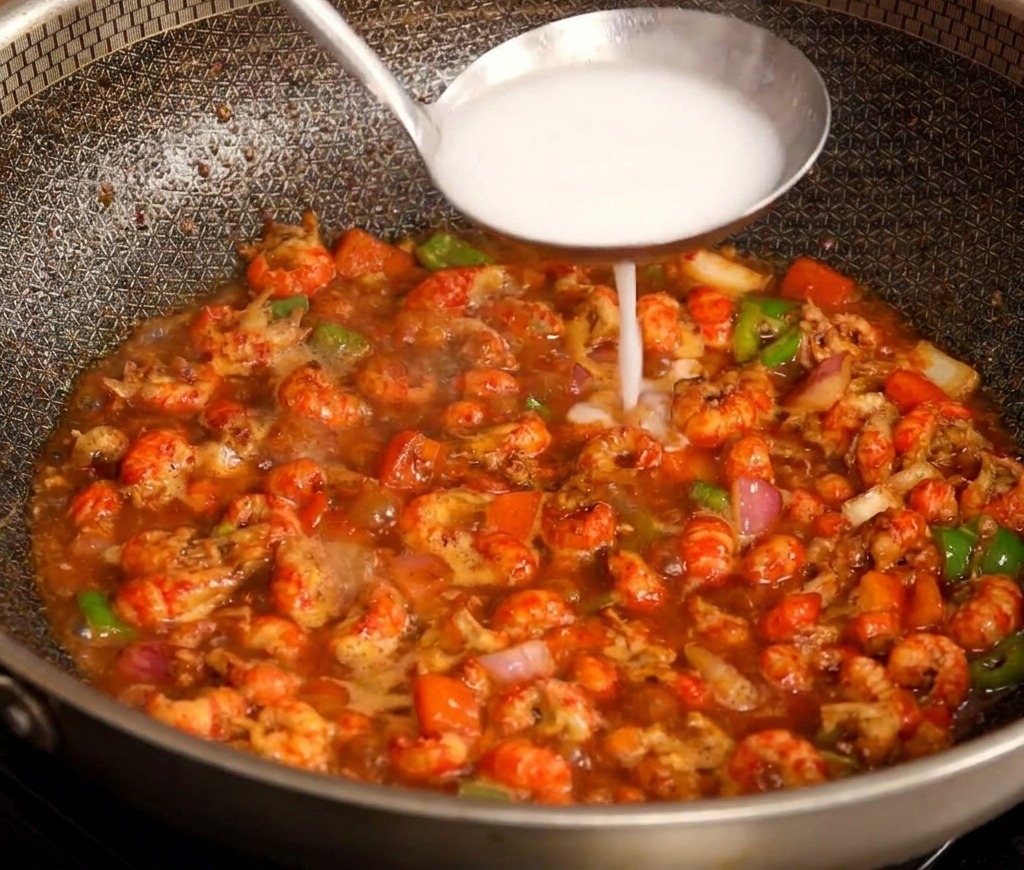 A stainless steel ladle pouring a white starch mixture into the center of a simmering pan of red, spicy crayfish.