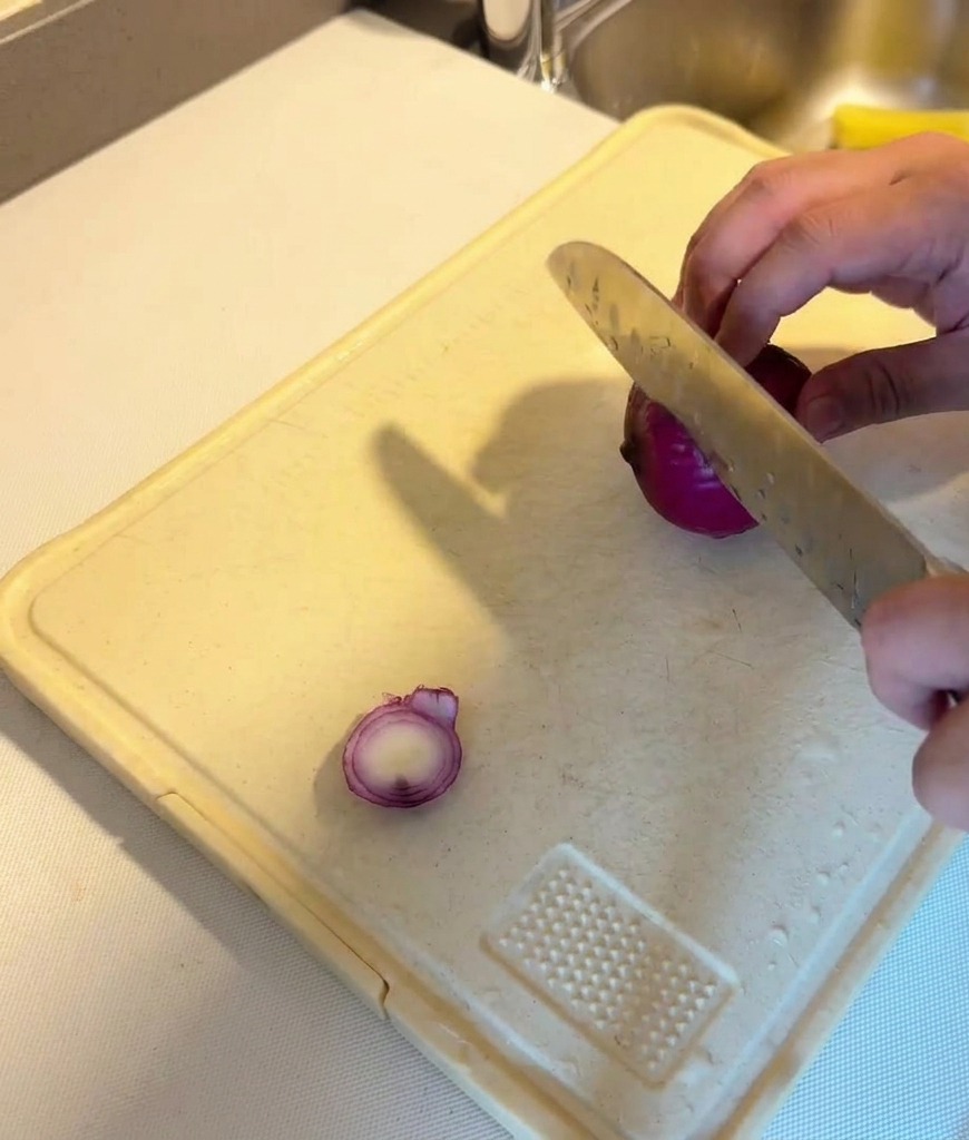A person using a silver knife to slice a purple red onion into rounds on a white plastic cutting board.