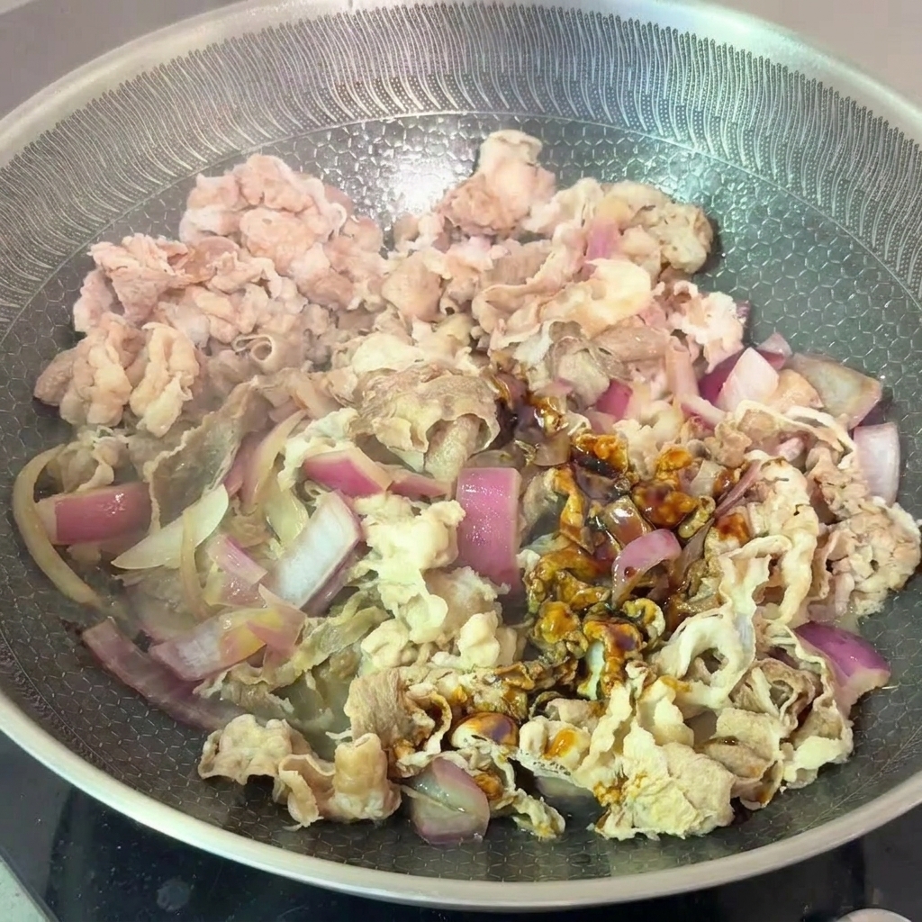 Blanched beef slices and red onions being tossed with a dark soy-based sauce in a frying pan.