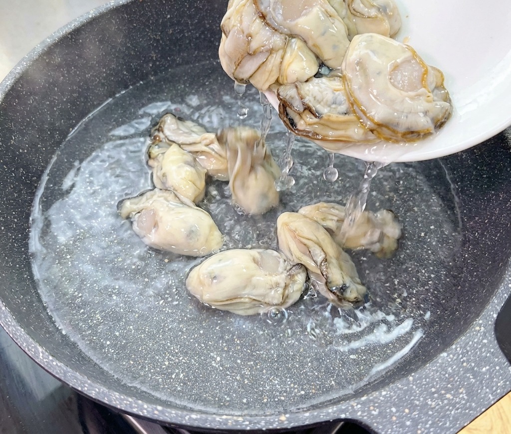 Raw, fresh oysters are sliding from a white plate into a wok containing cold water.