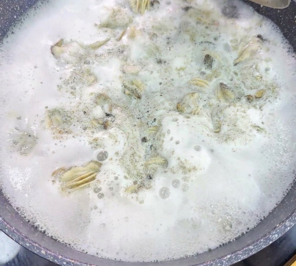 A metal strainer lifts poached oysters out of a pot of vigorously boiling, foamy water.