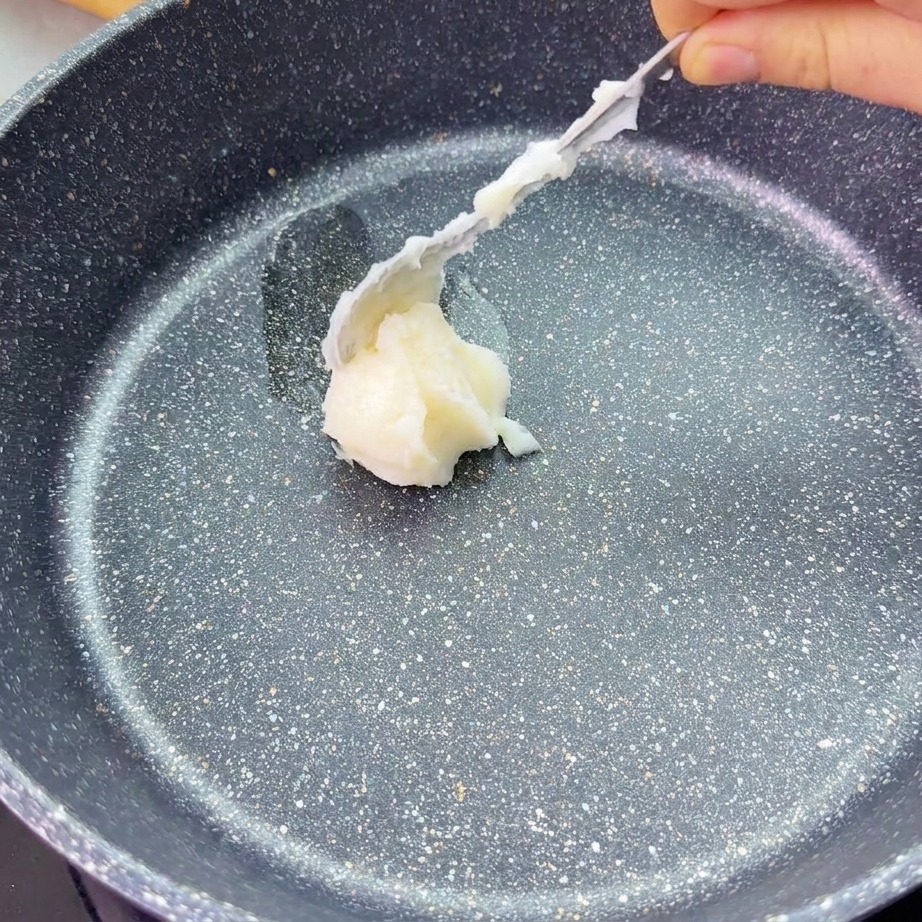 A spoon places a dollop of white lard into the center of a dark, stone-finish frying pan.