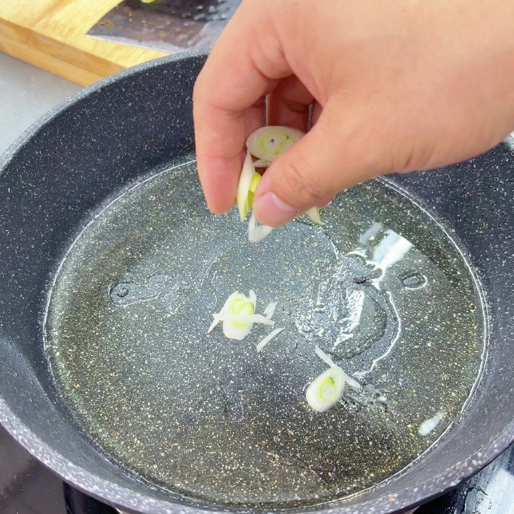 Slices of green and white scallions being stir-fried in a shallow layer of oil in a dark granite-textured pan.