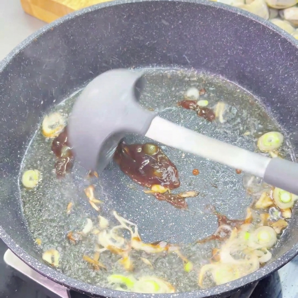 A dark, thick oyster sauce being stirred into sautéed scallions in a frying pan using a grey silicone spatula.