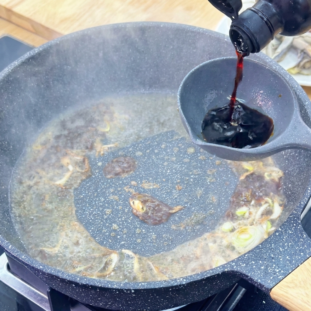 Light soy sauce being poured from a dark glass bottle into a grey ladle over a bubbling sauce in a pan.
