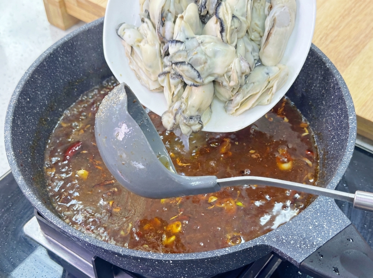 A plate of large, plump, blanched oysters being slid into a bubbling brown sauce in a granite-finish skillet.