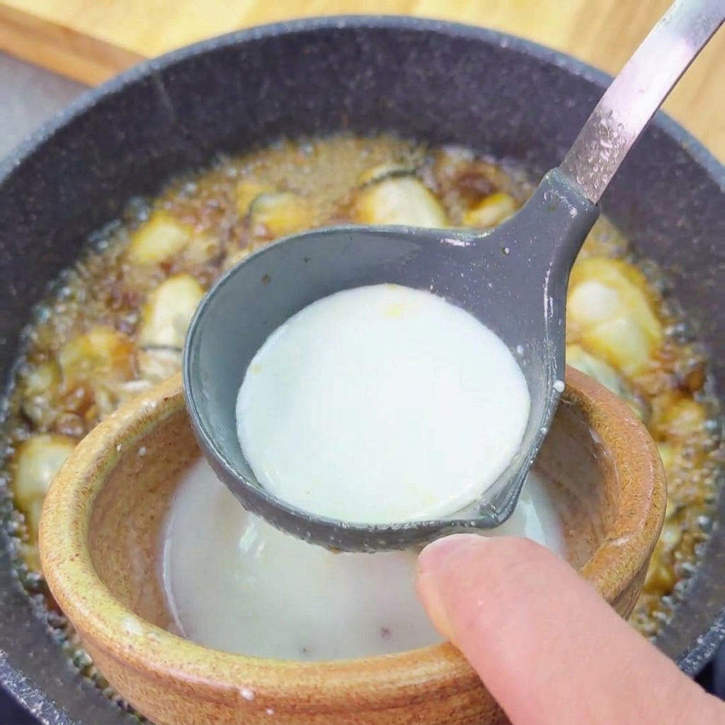 A milky white potato starch slurry being poured from a small bowl into a grey ladle over a simmering oyster dish.