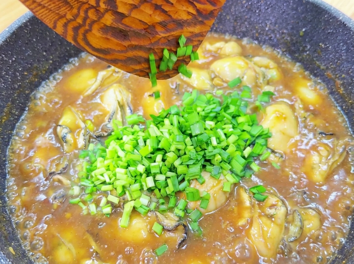 Freshly chopped green chives being dropped from a wooden spatula into a pan of simmering oysters in a thick, savory brown sauce.