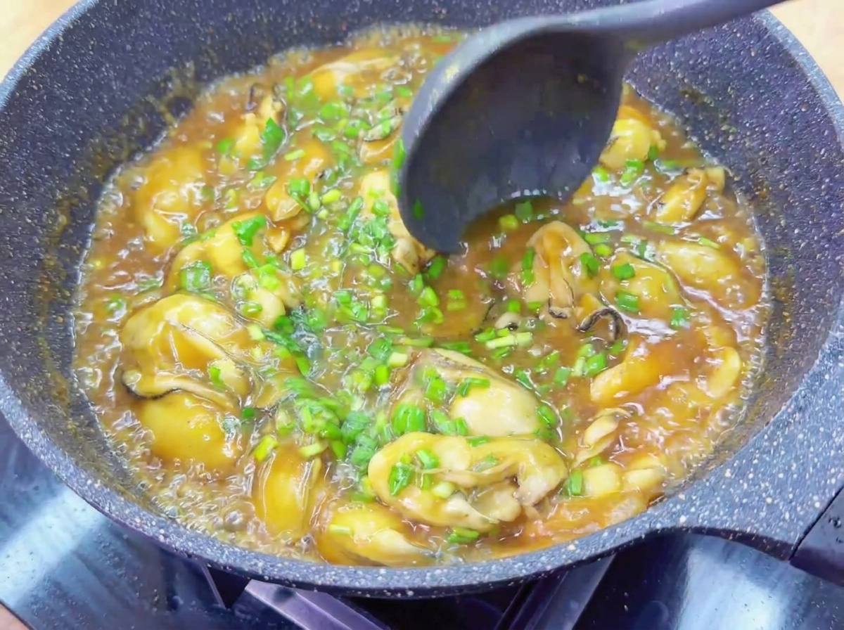 A dark ladle stirring together whole oysters and green chives in a pan filled with bubbling, glossy brown sauce.