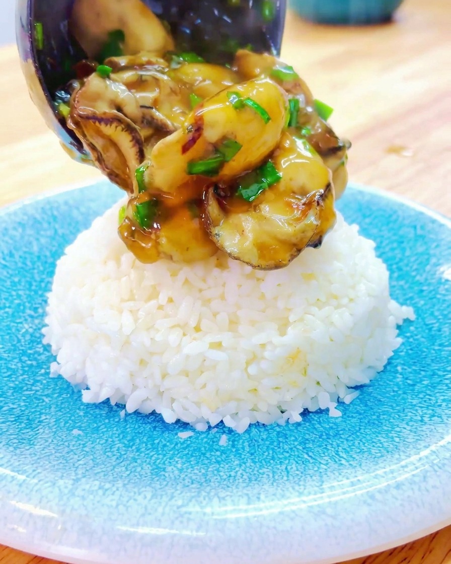 A ladle pouring thick, savory gravy and whole oysters onto a perfectly shaped mound of white rice served on a blue ceramic plate.