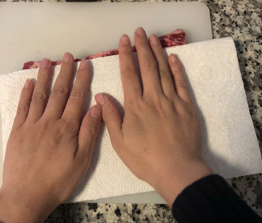 Hands using a white paper towel to absorb moisture from raw beef short ribs laid out on a white cutting board.