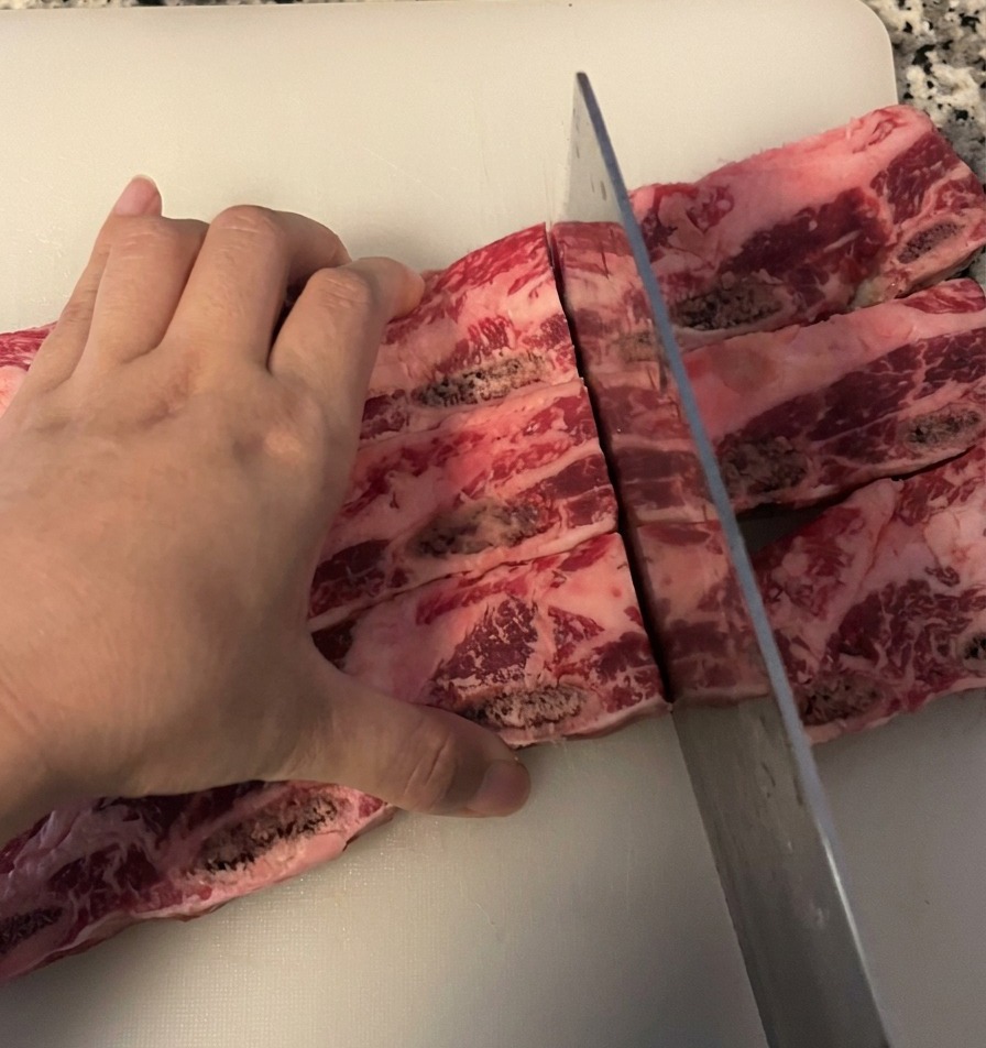 A person slicing a slab of raw beef short ribs into individual bone in pieces using a large steel chef knife on a white cutting board.