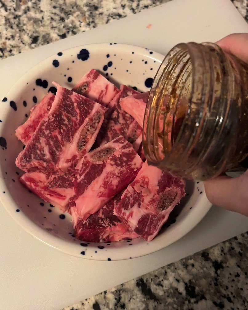 Dark glossy Korean BBQ sauce being poured from a glass jar onto a pile of raw sliced beef short ribs in a blue speckled white bowl.