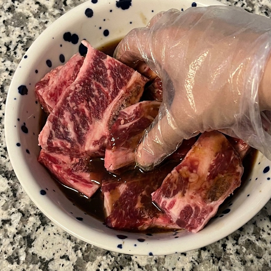 A hand wearing a clear plastic glove mixing raw beef short ribs with a dark marinade in a bowl to ensure even coating.