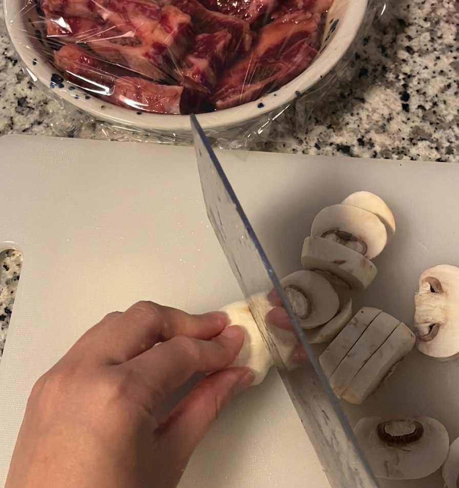 A hand holding a white button mushroom while slicing it into thick rounds with a kitchen knife on a white cutting board.