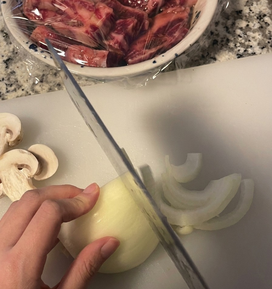 A person slicing a white onion into thin half-moon segments on a white cutting board using a kitchen knife.