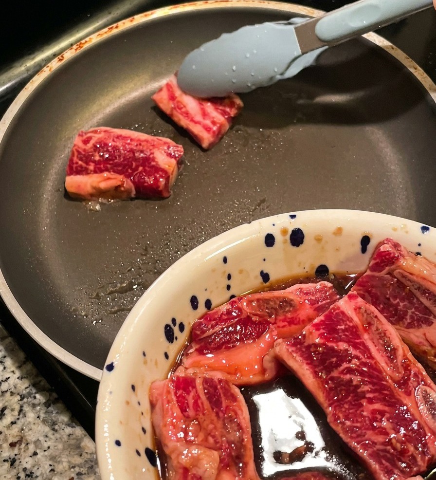 Tongs placing thick slices of marinated raw beef short ribs into a hot frying pan, with a bowl of more meat visible in the foreground.
