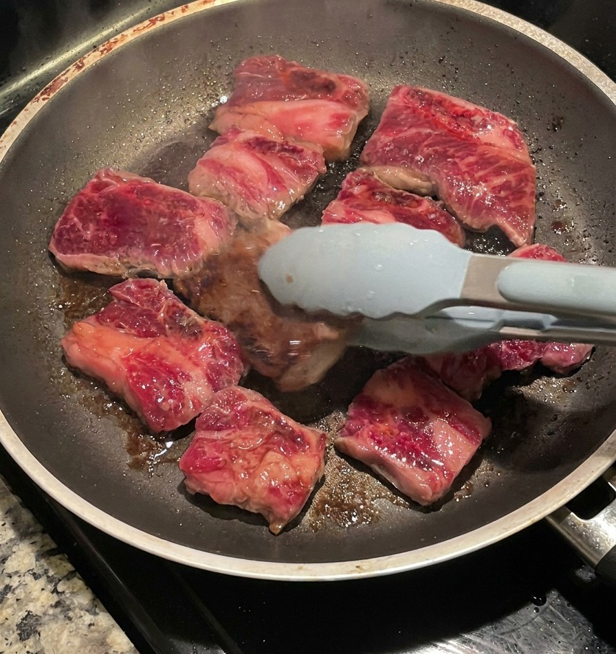 Using blue-tipped tongs to flip seared beef short ribs in a pan, revealing a dark, caramelized crust on the cooked side.