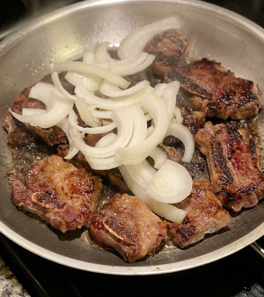 A pile of raw, sliced white onions sitting on top of browned beef short ribs in a stainless steel pan.