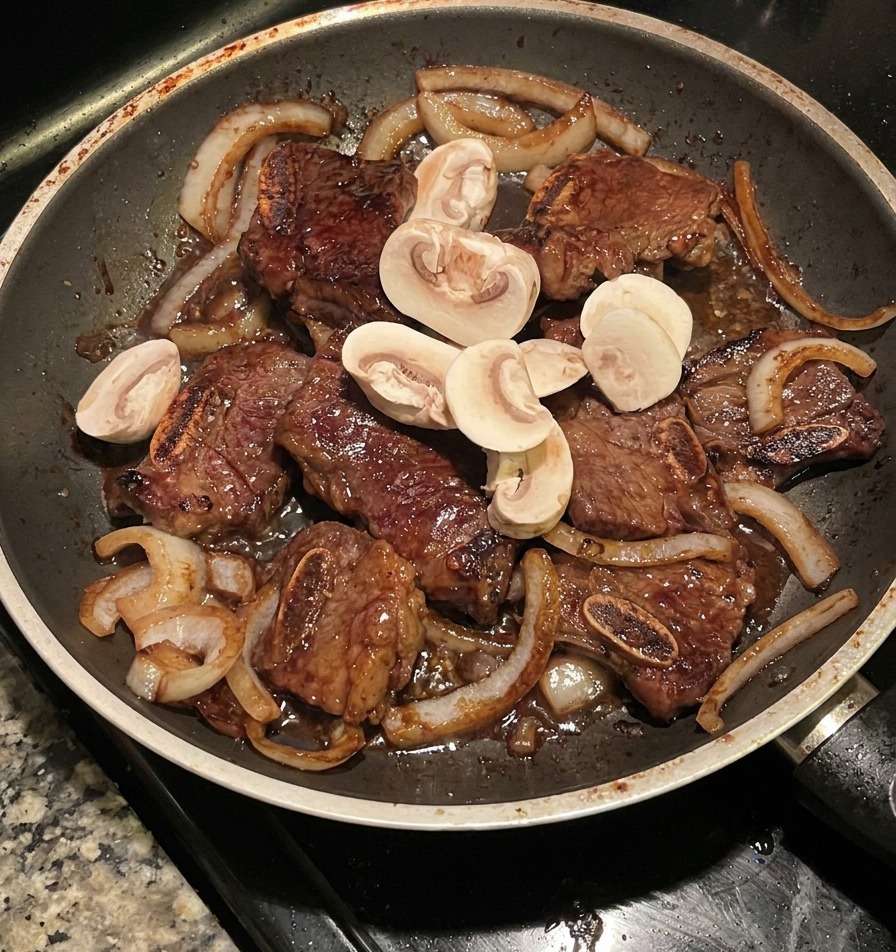 Sliced mushrooms being stir-fried with beef ribs and caramelized onions in a dark frying pan.