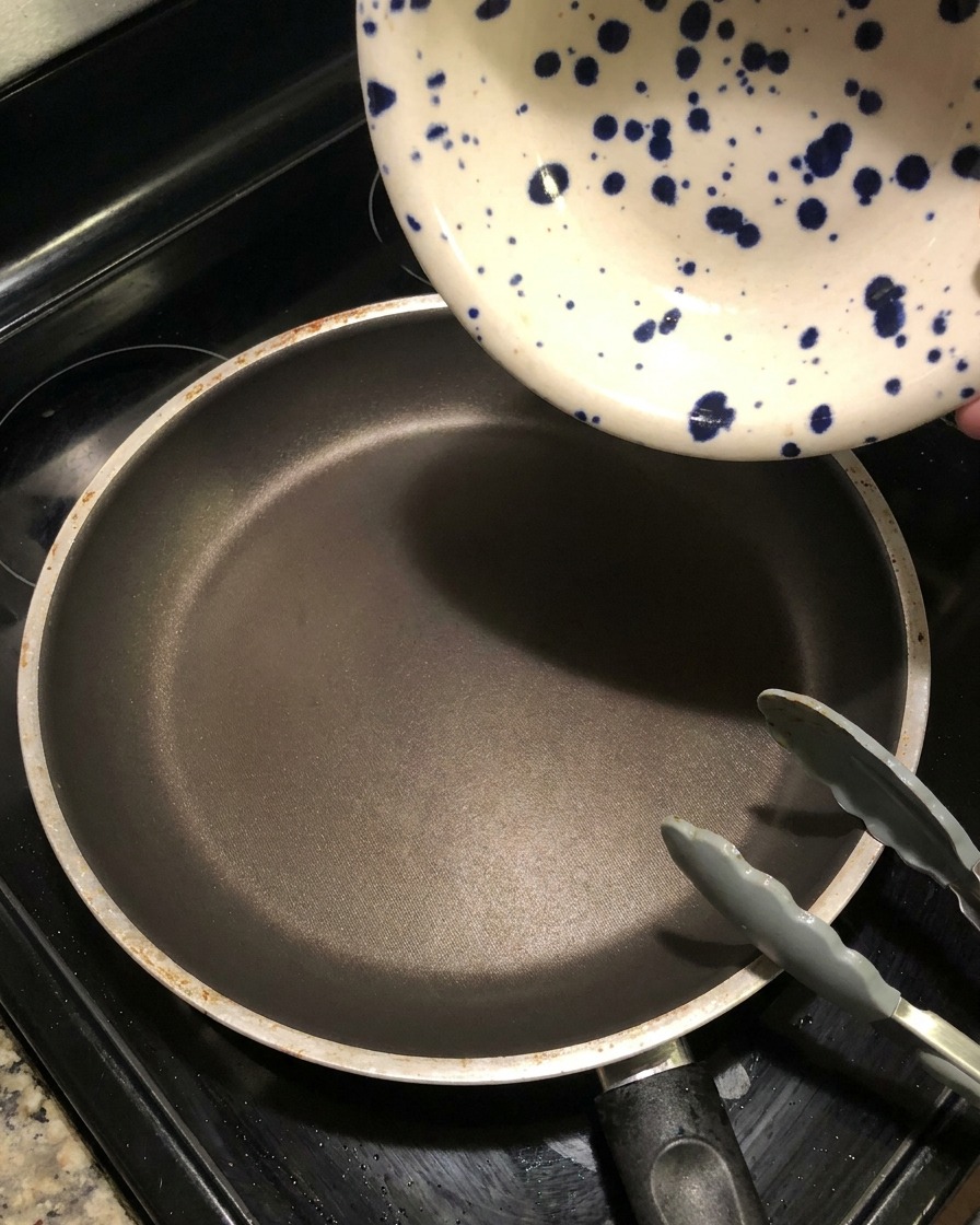 A blue-speckled ceramic bowl pouring dark marinade liquid into a hot, empty frying pan to create a sauce reduction.