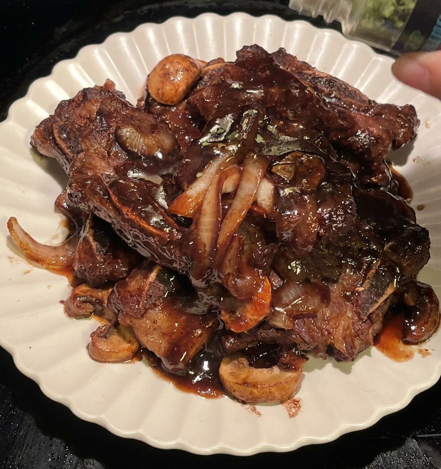 Finely chopped green parsley being sprinkled from a spice jar onto the plated BBQ short ribs and mushrooms.